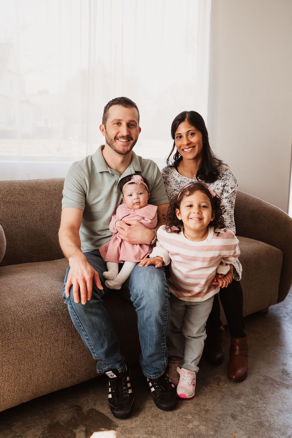 Happy family of four posing on a couch, parents smiling with a toddler girl and baby in pink dress.