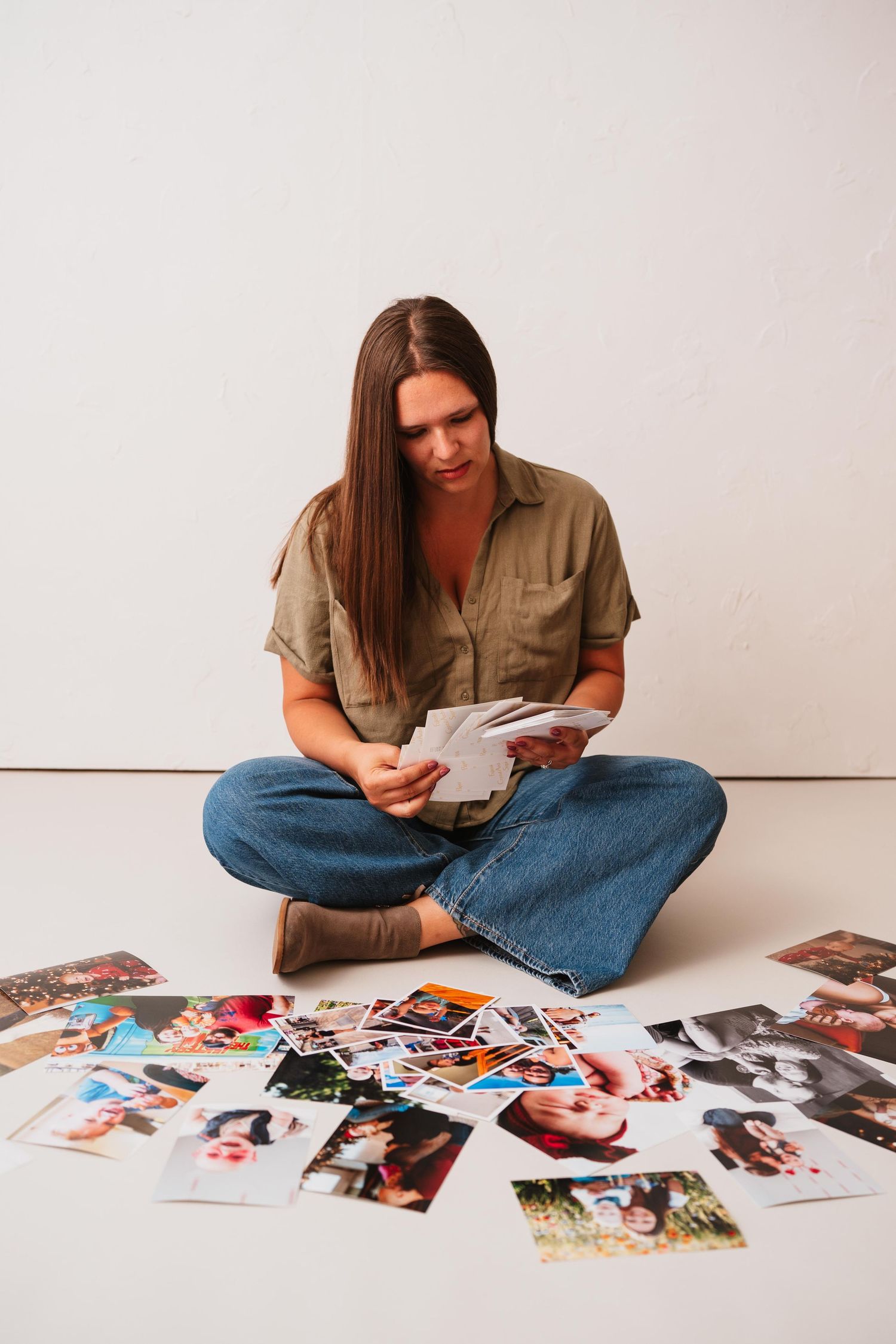 Woman with long brown hair sits cross-legged on a white studio floor, sorting printed photographs spread out around her.