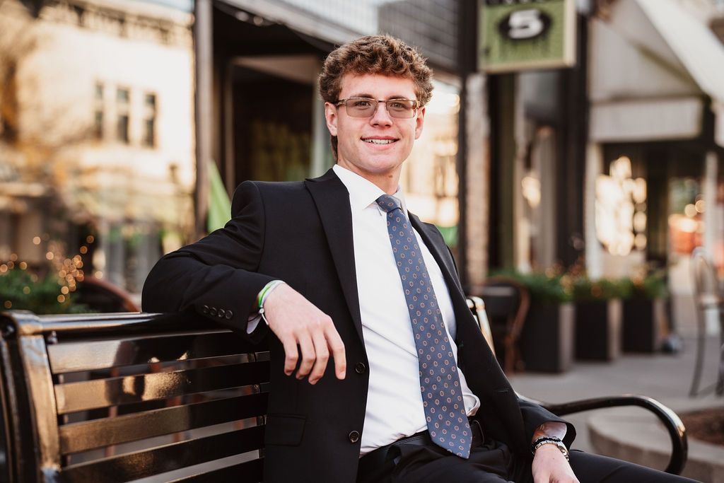 Young professional man in suit and tie sitting on bench in urban setting, smiling confidently outdoors.