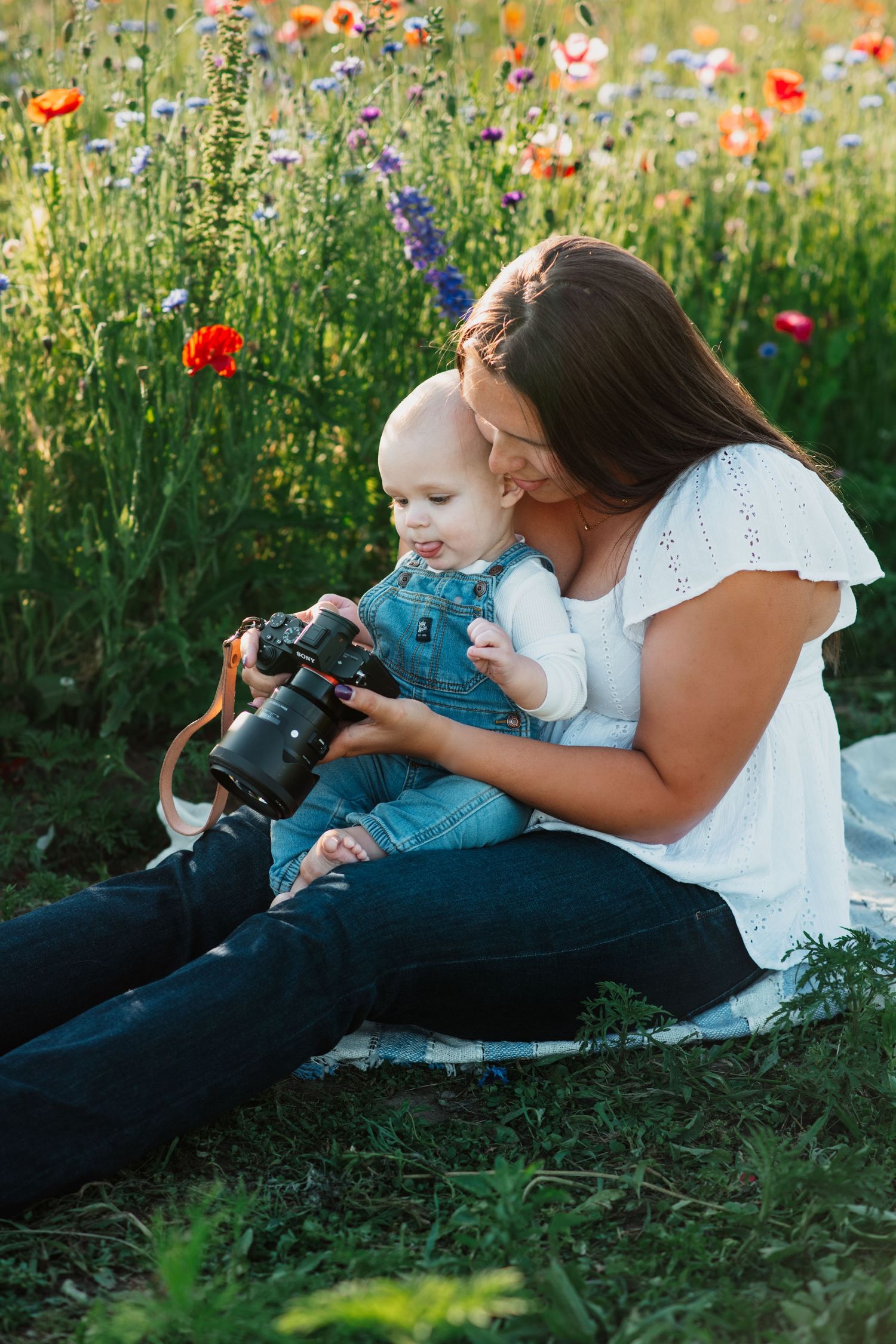 Mother and baby sitting in a wildflower field, looking at a camera together on a sunny day.