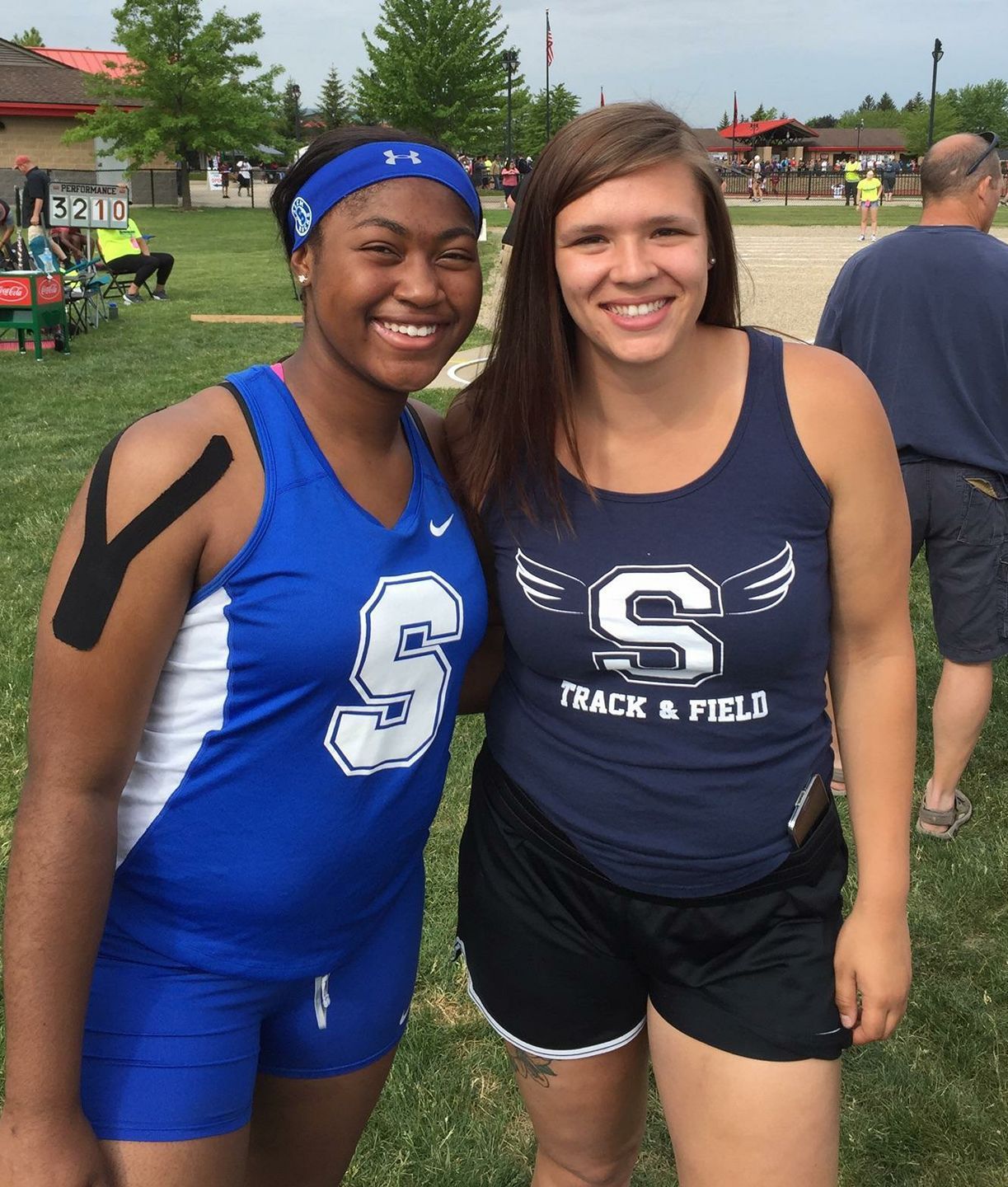 Two female track and field athletes in blue uniforms smiling together at an outdoor meet.