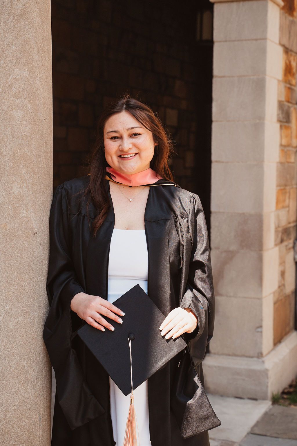 Smiling graduate in black cap and gown holds mortarboard at University of Michigan building.