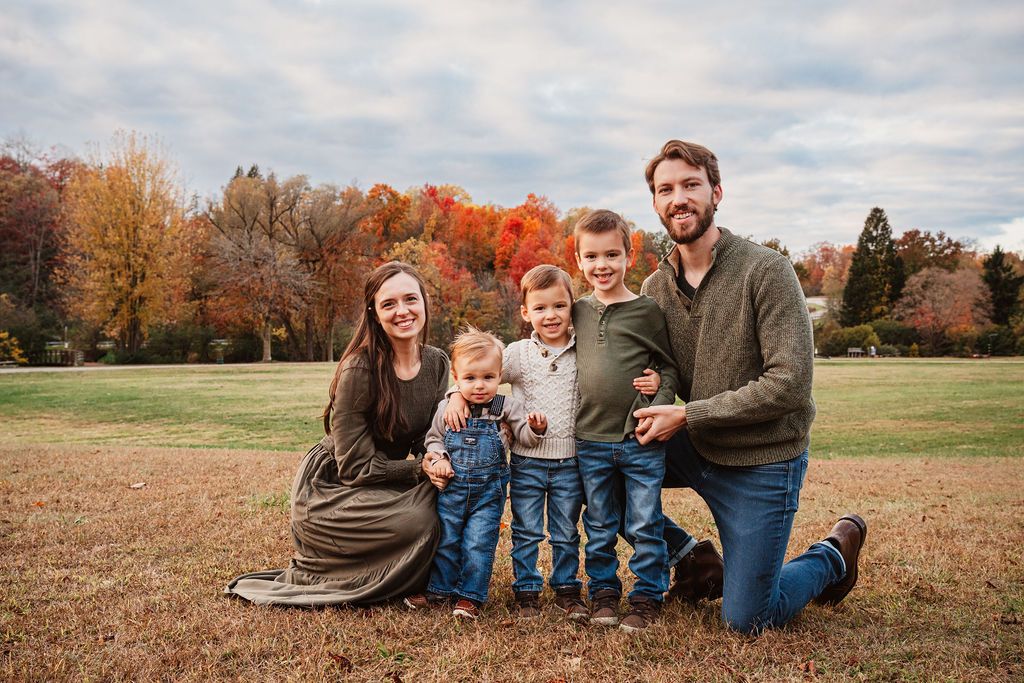 Family of five posing outdoors in a fall park with colorful autumn foliage in the background.