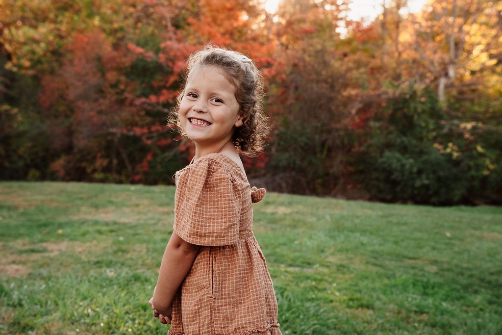 A smiling young girl in an orange gingham dress stands on green grass with colorful autumn trees in the background.