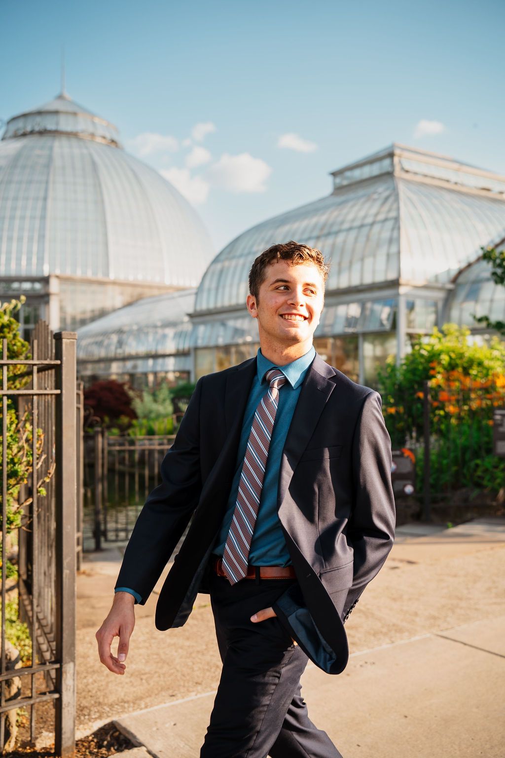 Young man in dark suit and striped tie smiling while walking outside a glass greenhouse conservatory.