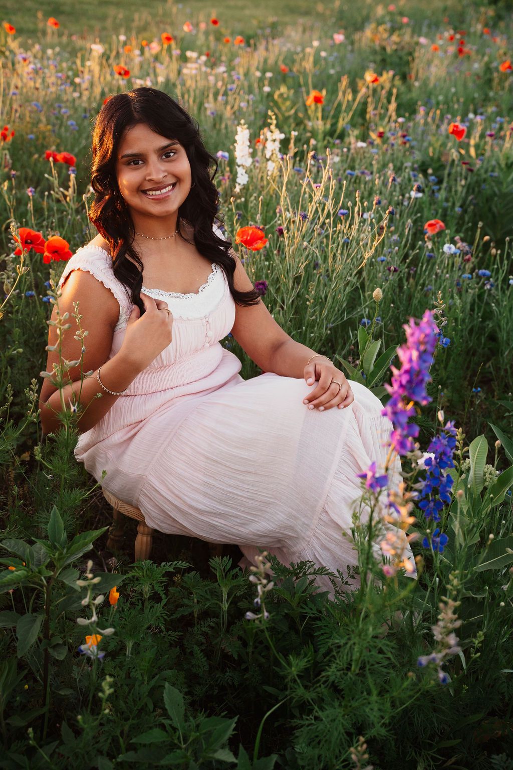 Young woman in pink dress sitting among colorful wildflowers, smiling outdoors in a vibrant garden setting.