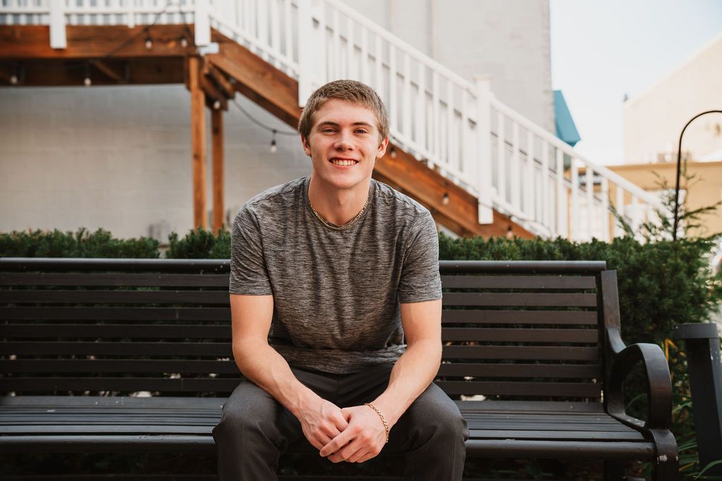 Young man smiling while sitting on a dark wooden bench outdoors near stairs and green shrubs.