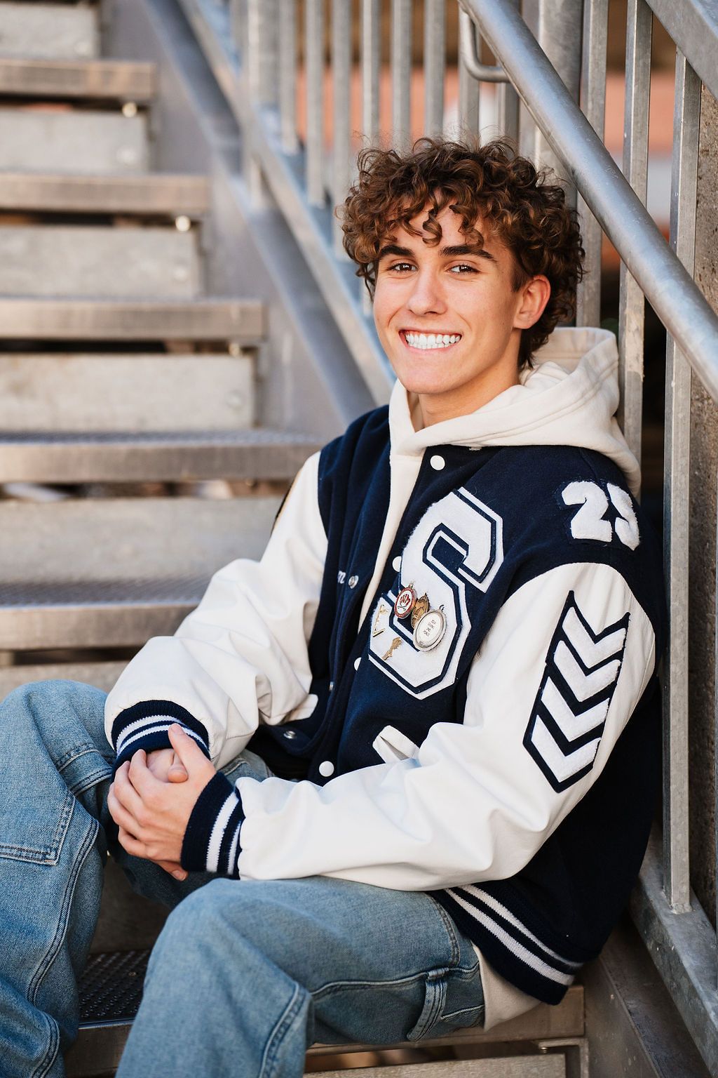 Smiling teen in varsity letterman jacket sitting on bleacher steps for senior portrait photo.