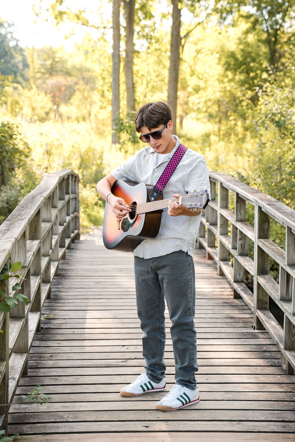 Young man in sunglasses playing acoustic guitar on a wooden bridge surrounded by lush green trees in natural sunlight.