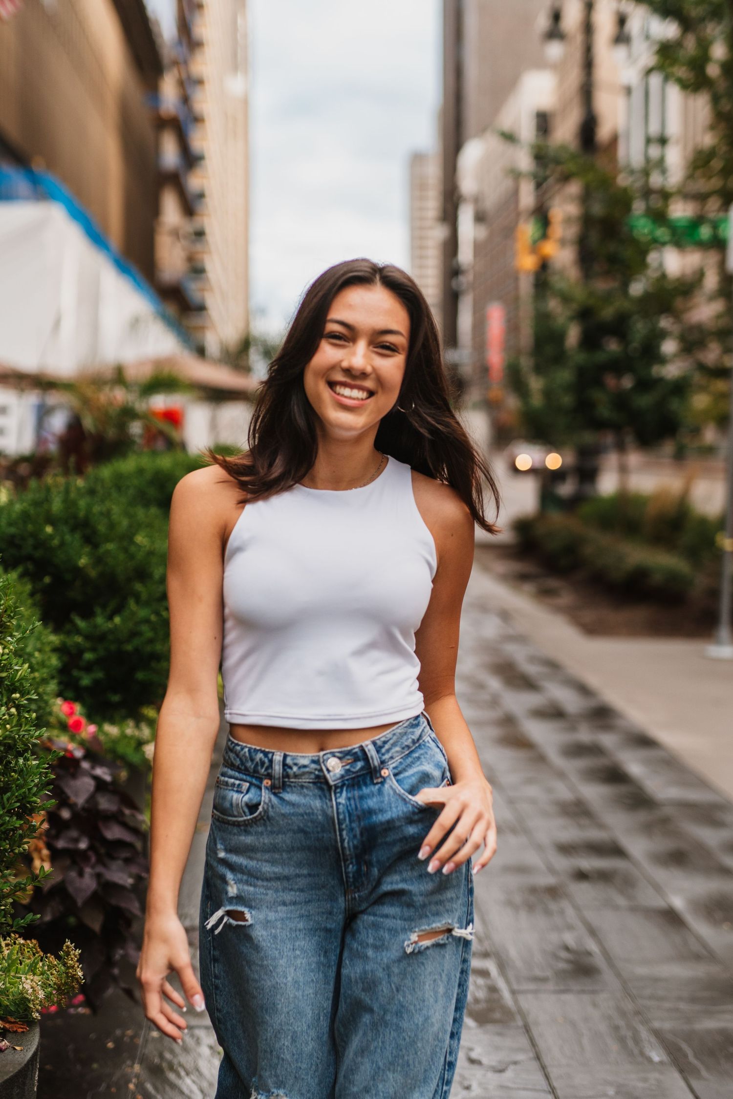Smiling young woman in white crop top and ripped jeans walking on a city sidewalk with urban buildings in background.