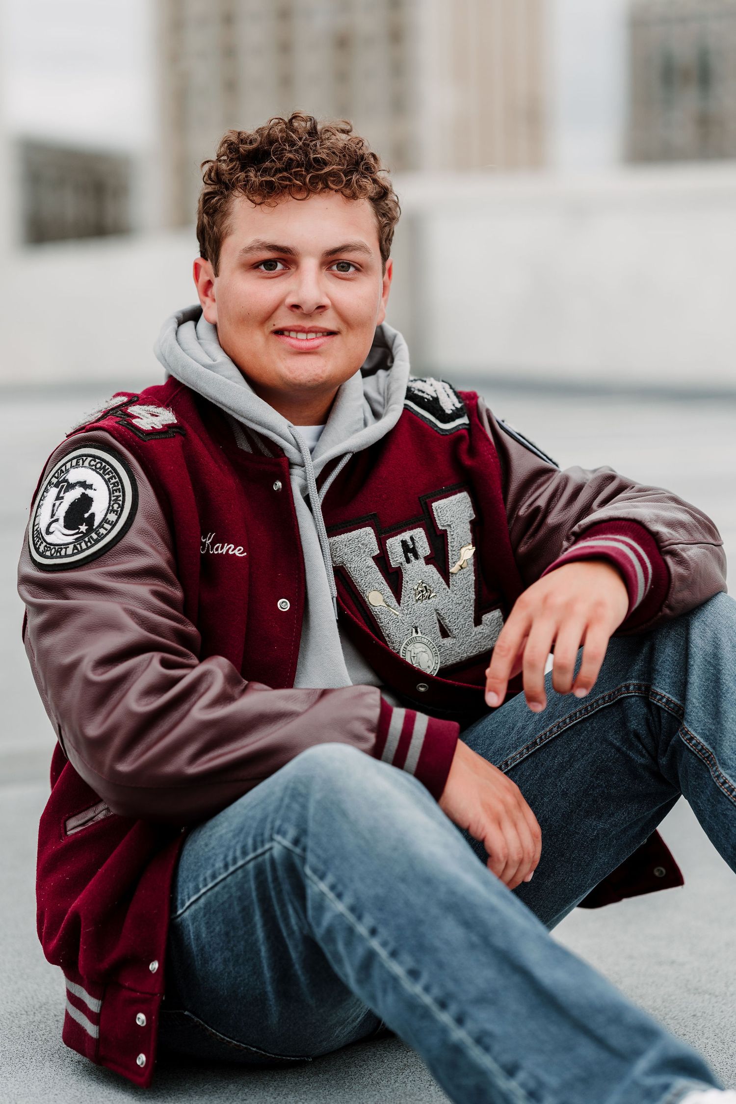 Smiling young man in a maroon varsity letterman jacket sits casually on the ground outdoors.