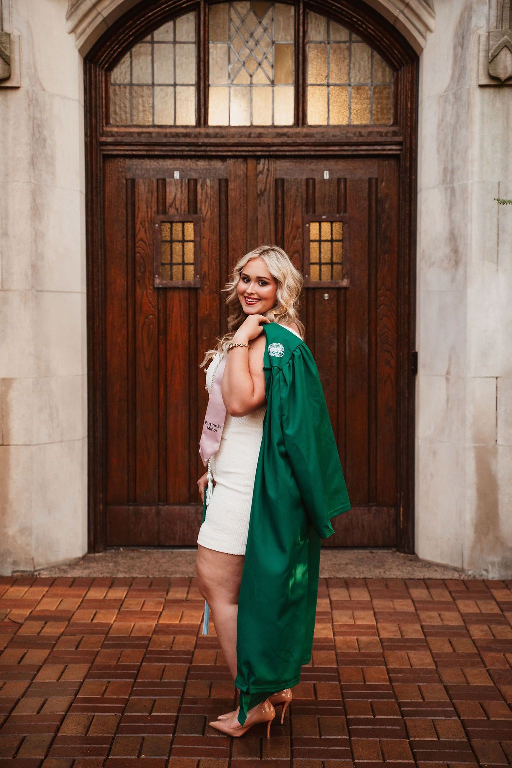 Smiling blonde graduate in green gown and white dress poses in front of ornate wooden doors on brick steps.