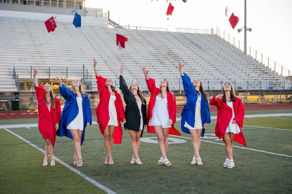 Graduates in red and blue gowns stand in formation on a football field during commencement ceremony.