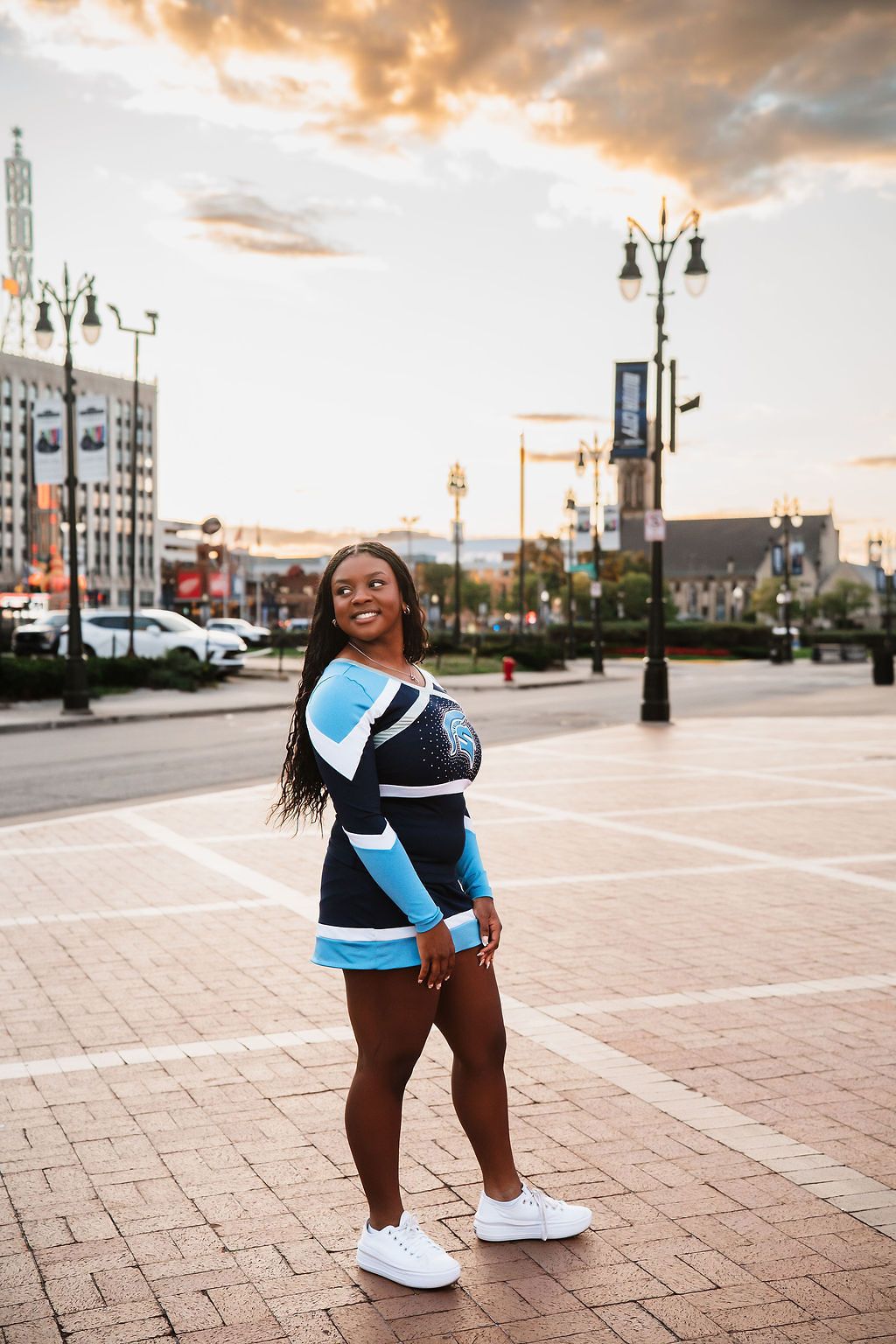 Cheerleader in blue and black uniform posing on an urban plaza at sunset with city lights in the background.