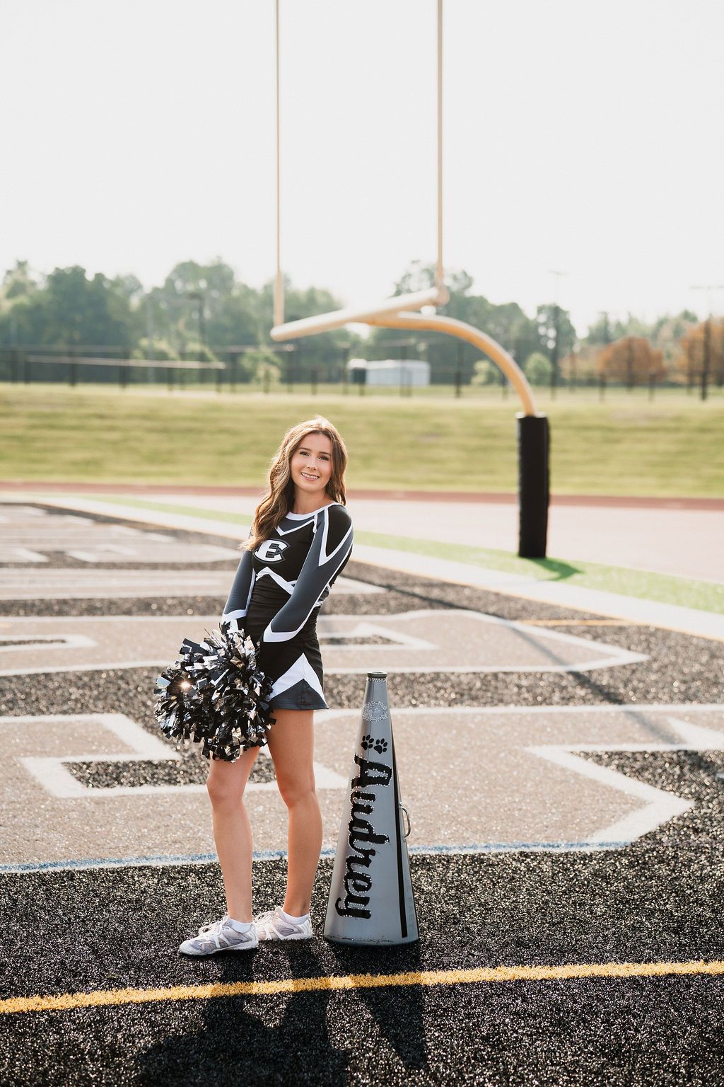 Cheerleader in black and white uniform poses on football field track holding pom poms next to a personalized megaphone.