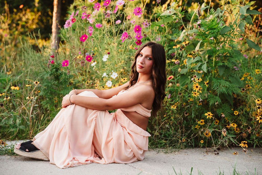 Young woman in pink dress sitting among colorful wildflowers in a garden for senior portrait.