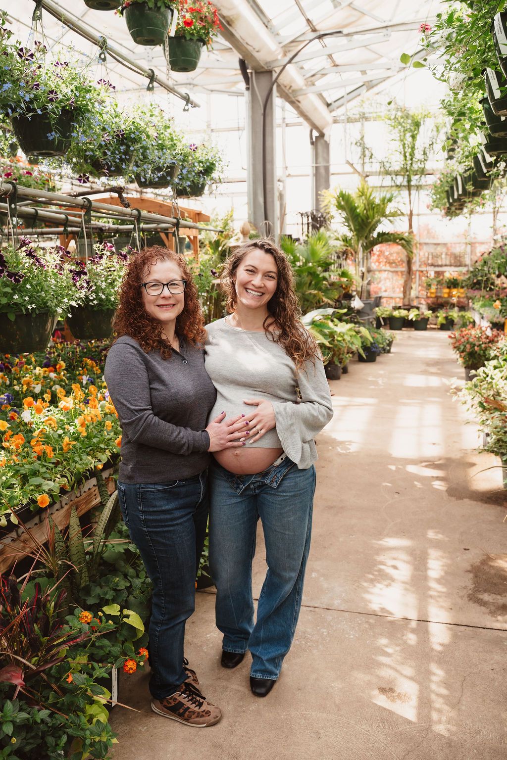 Two women pose among colorful flowers in a greenhouse, one pregnant cradling her baby bump.