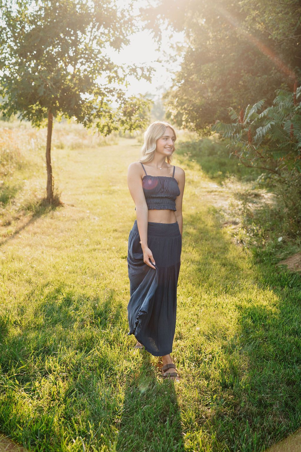A maternity photoshoot in a sunlit meadow with golden light filtering through trees during sunset.