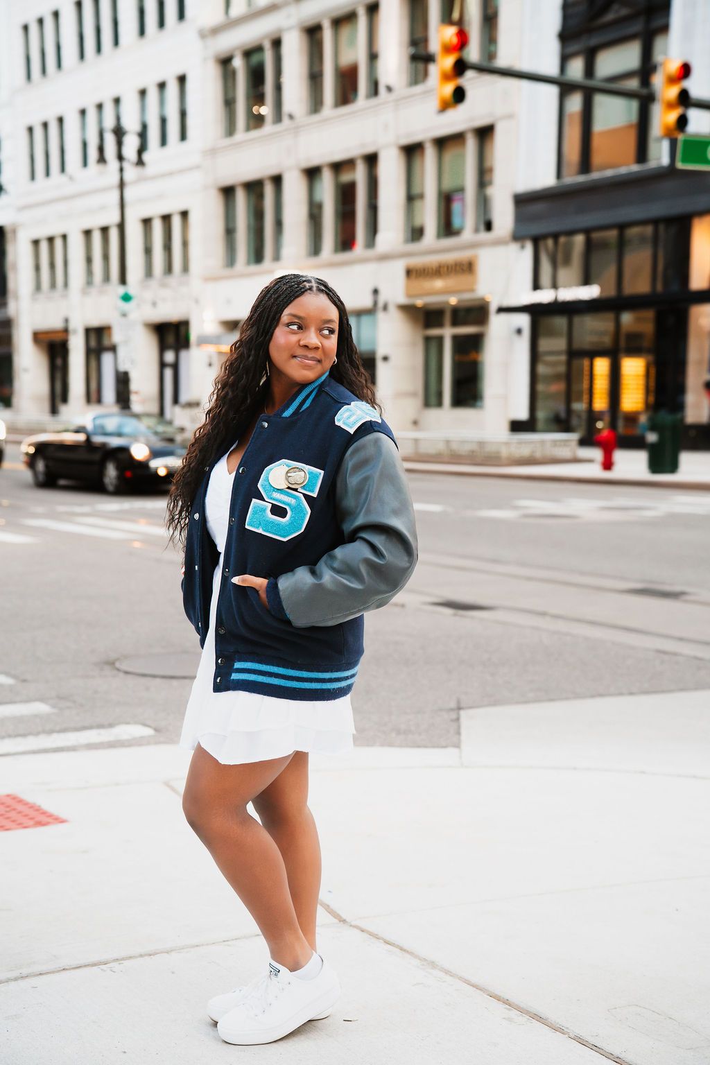 A person wearing a varsity letterman jacket and white dress poses on a city street corner with white sneakers.