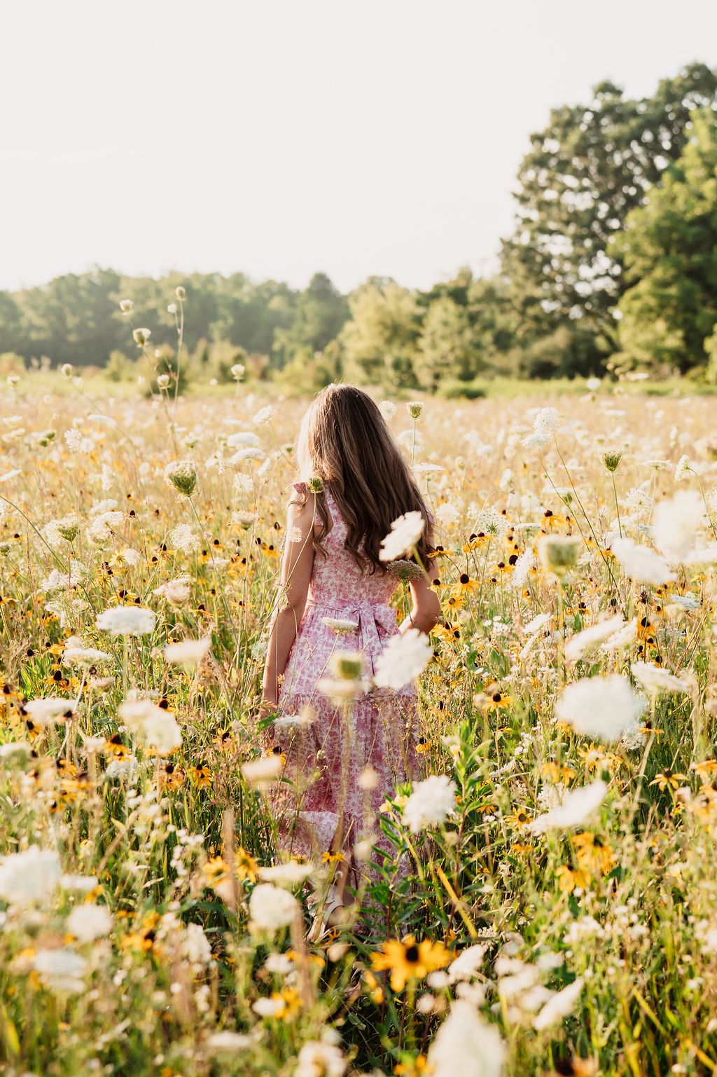 Woman in floral dress walking through dreamy summer meadow filled with white cosmos and wildflowers at sunset.