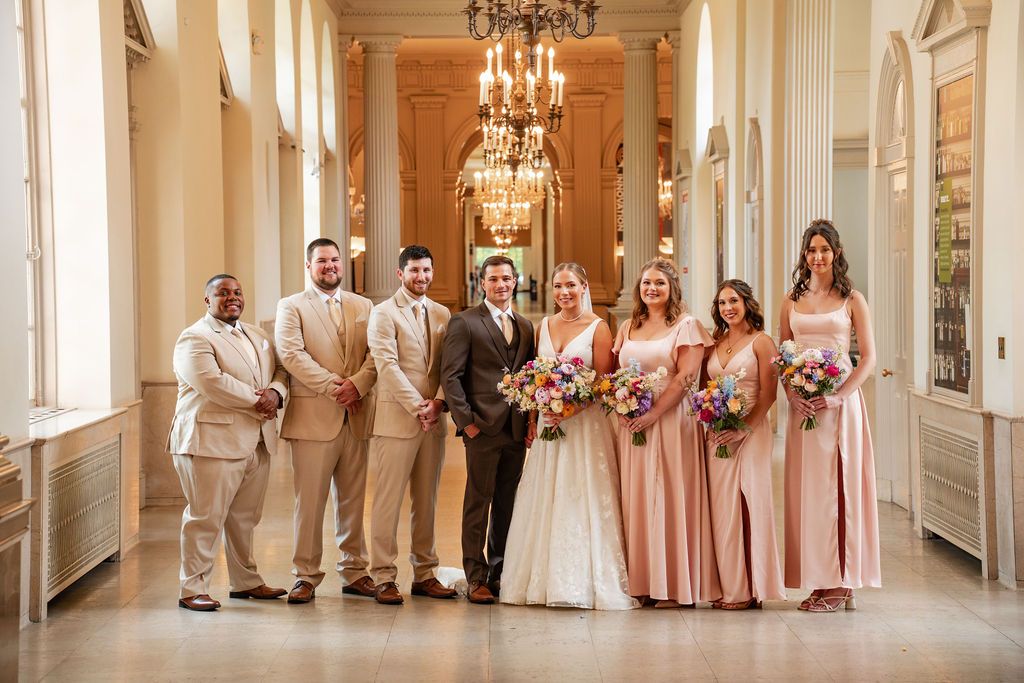 Wedding party lined up along elegant hallway with chandeliers and classic architecture.