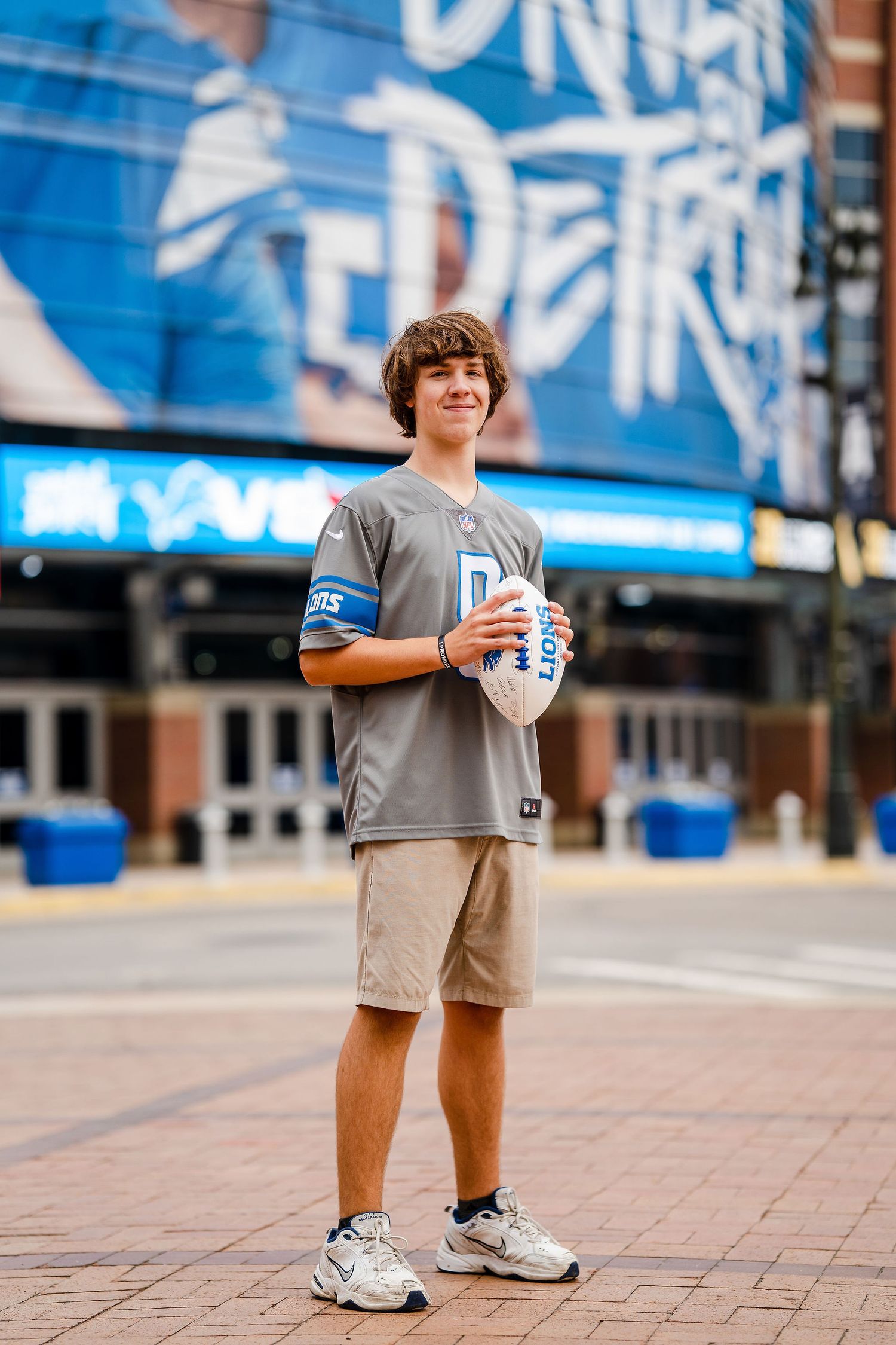 Young Detroit Lions fan holding a football outside Ford Field stadium in Detroit, wearing a gray Lions jersey.