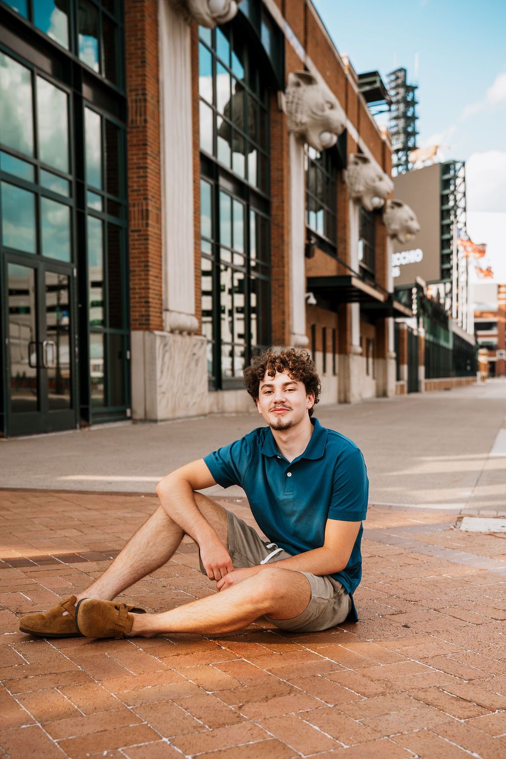 Young man with curly hair sits on brick sidewalk in blue polo shirt and khaki shorts outside urban building.