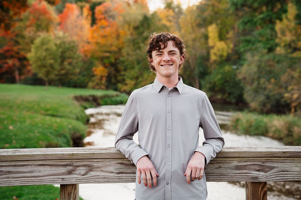 Smiling young man in gray button-up shirt leaning on wooden fence with colorful autumn trees behind him.