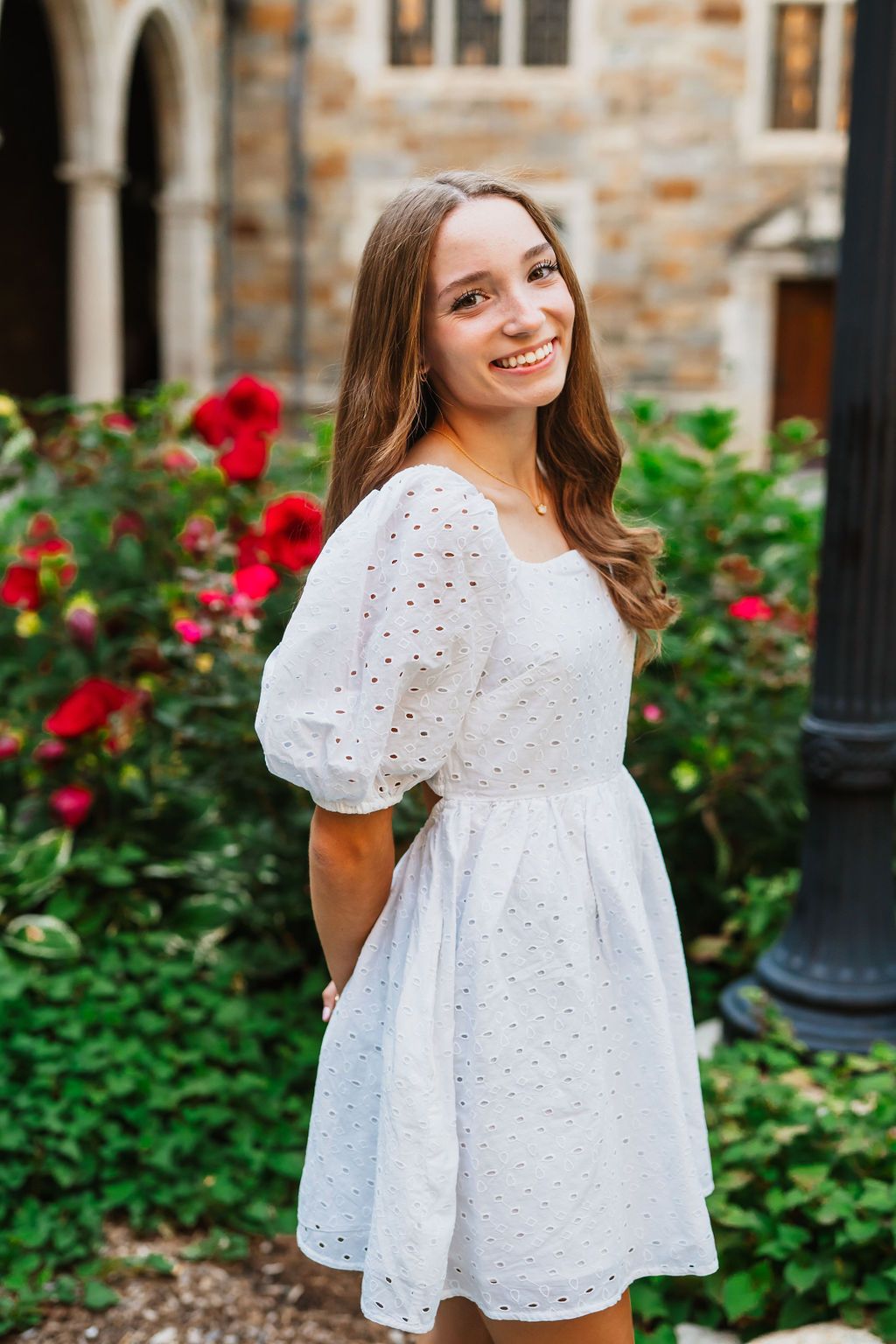 A person in a white eyelet summer dress poses in a garden with red roses and stone architecture in the background.