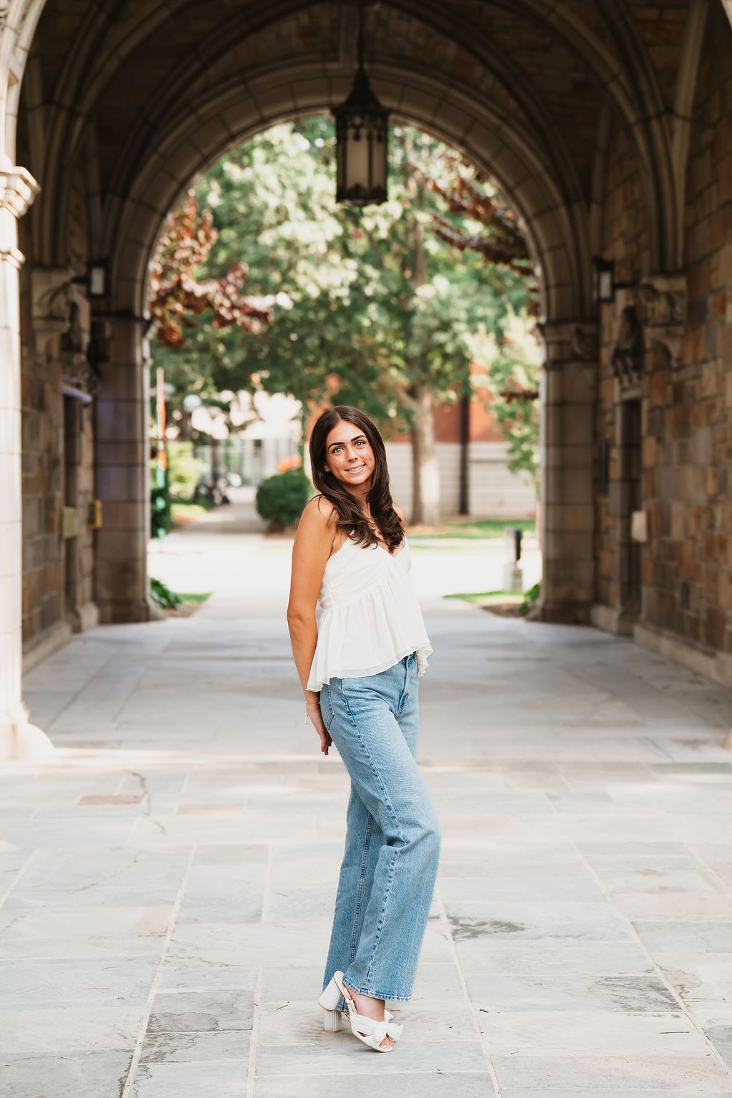A casual summer outfit featuring wide-leg jeans and a white top photographed under a stone archway on a university campus.