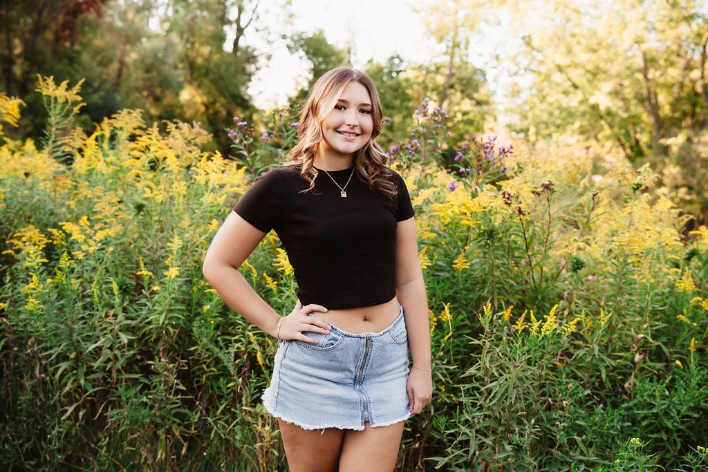A person poses in a black top and denim shorts against a backdrop of golden wildflowers during sunset.