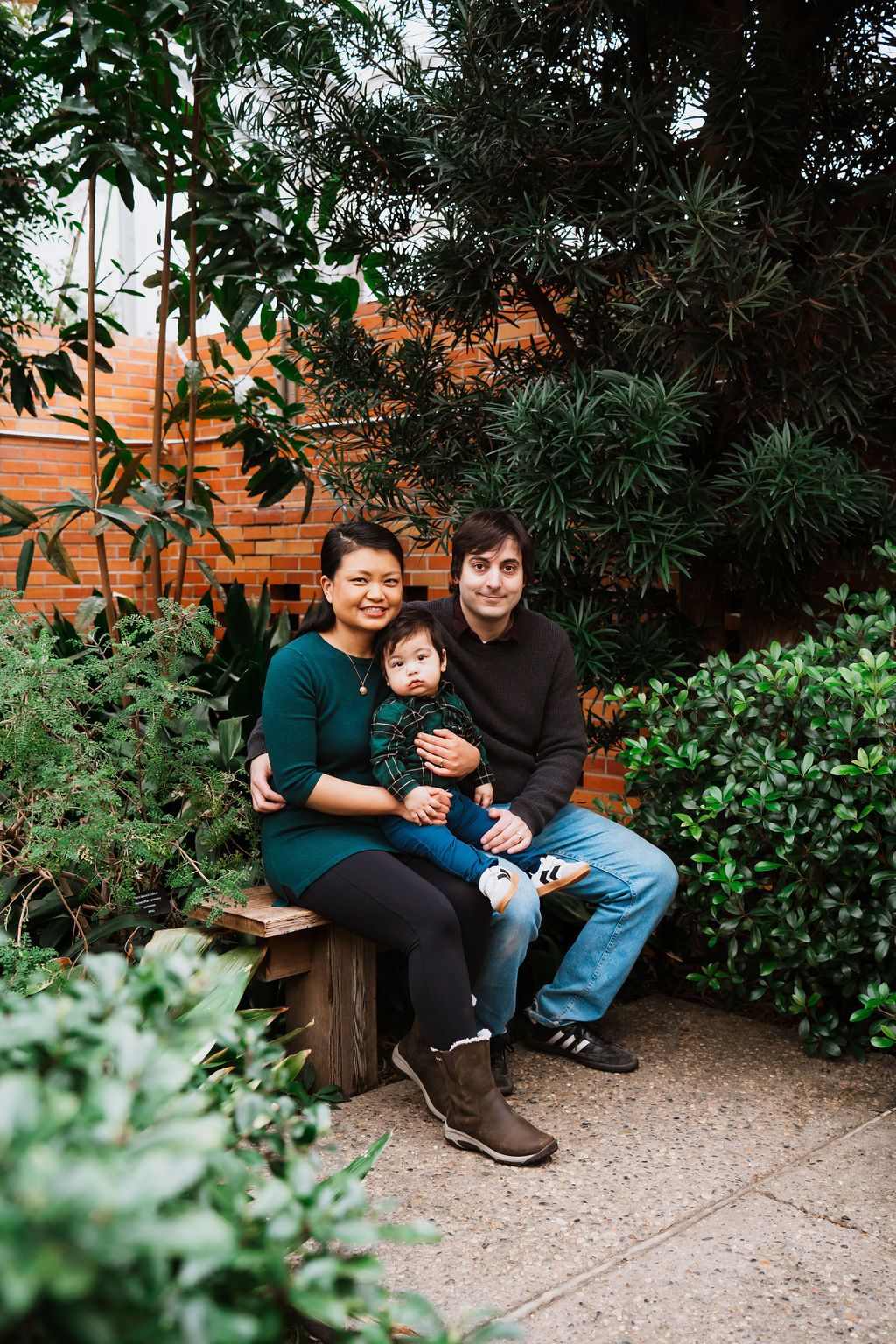 A happy family of three sits on a garden bench surrounded by lush greenery and a brick wall backdrop.