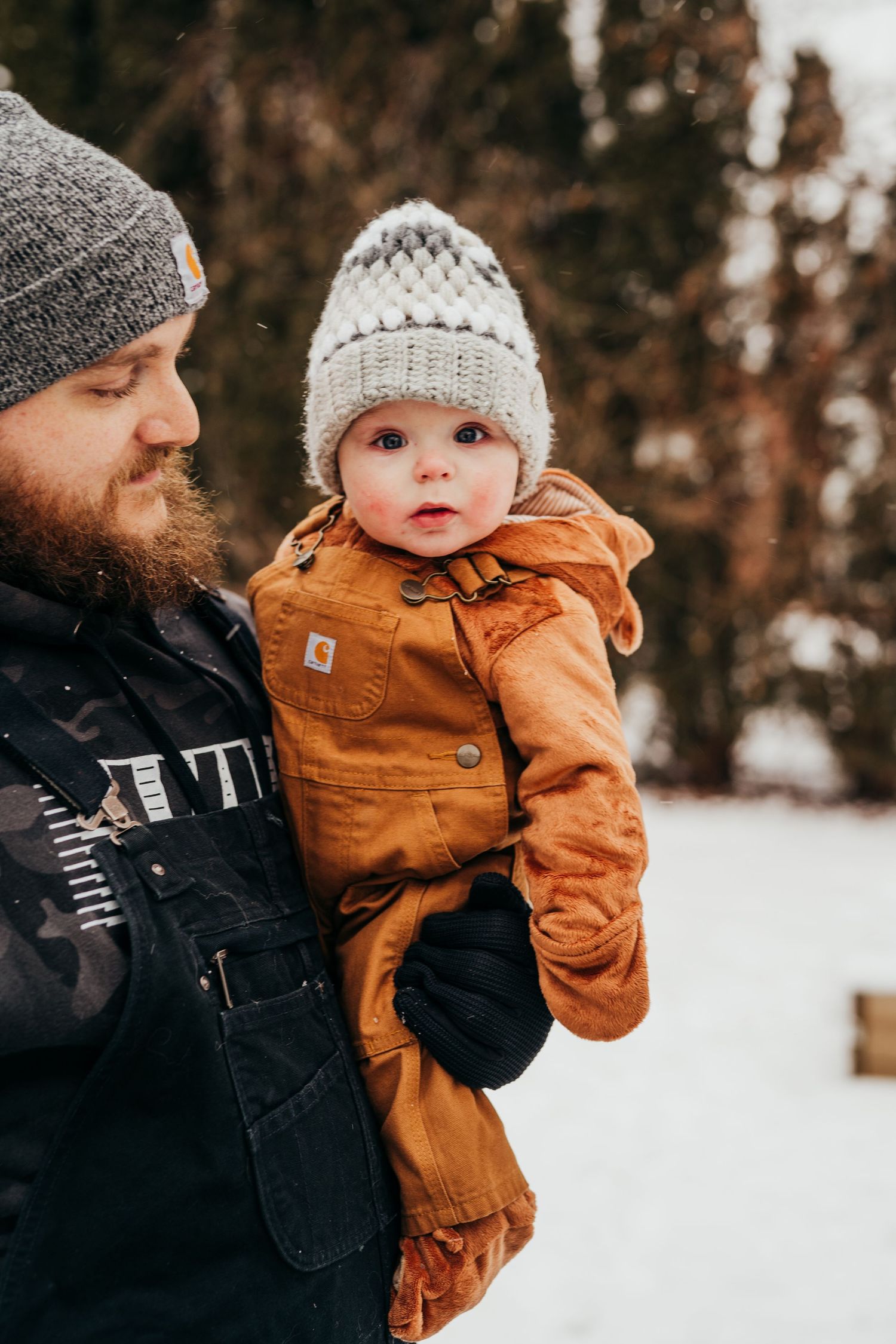 Bearded father holding baby in Carhartt outfit and knit hat in snowy winter outdoor setting.
