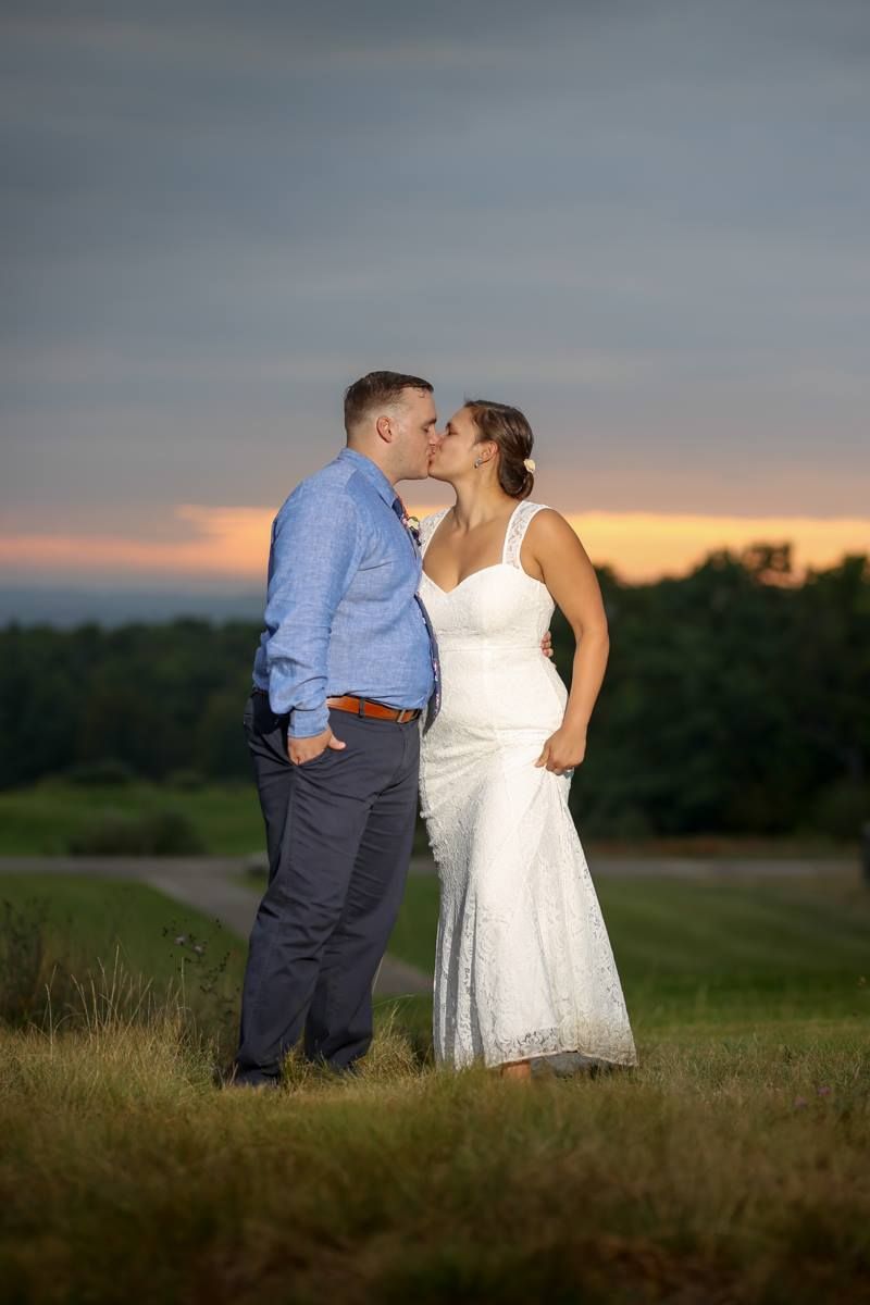 Couple sharing a romantic kiss in a field at sunset, bride in white lace gown and groom in blue shirt.