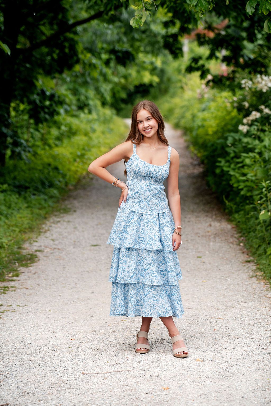 Young woman in blue floral tiered midi dress smiling on a tree-lined gravel path in summer.
