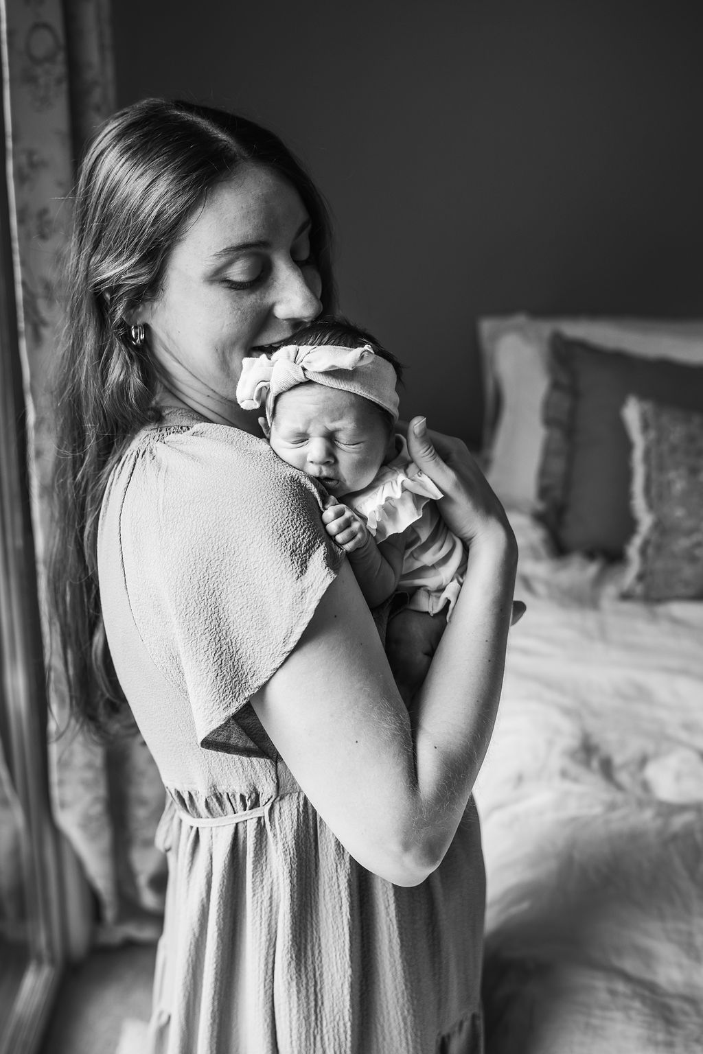 Black and white photo of a mother tenderly holding her newborn baby in a headband near a bedroom window.