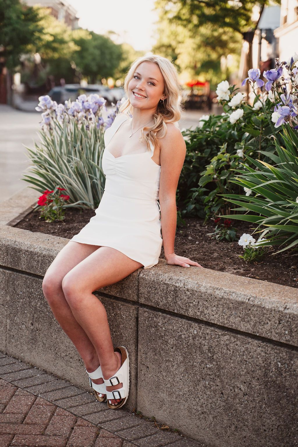 Young blonde woman in white mini dress and sandals sitting on a stone planter with purple flowers in an urban setting.