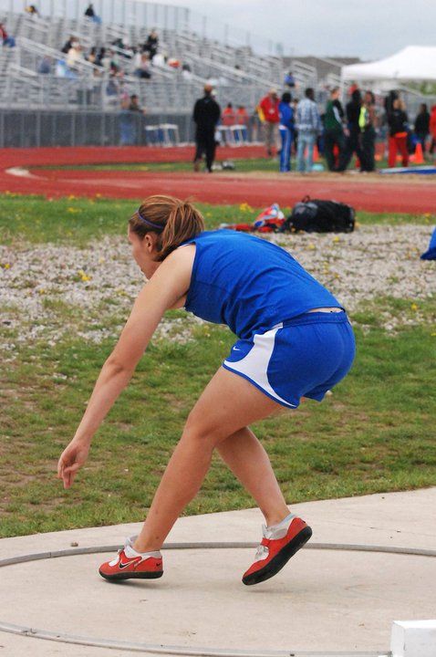 Female athlete in blue Nike uniform preparing to throw shot put at a track meet.
