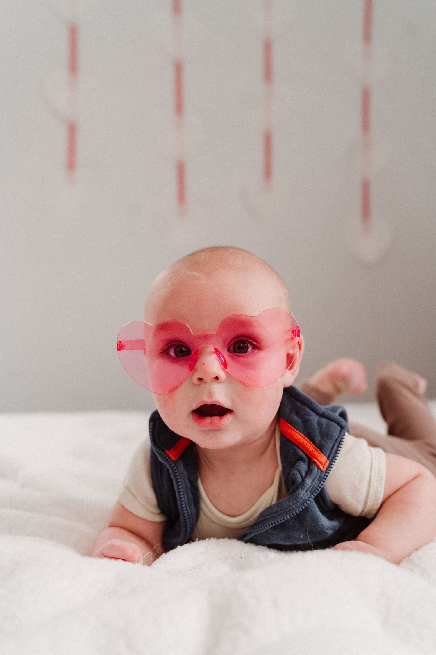 Adorable baby wearing oversized pink heart-shaped sunglasses lying on a white blanket with mouth open in surprise.