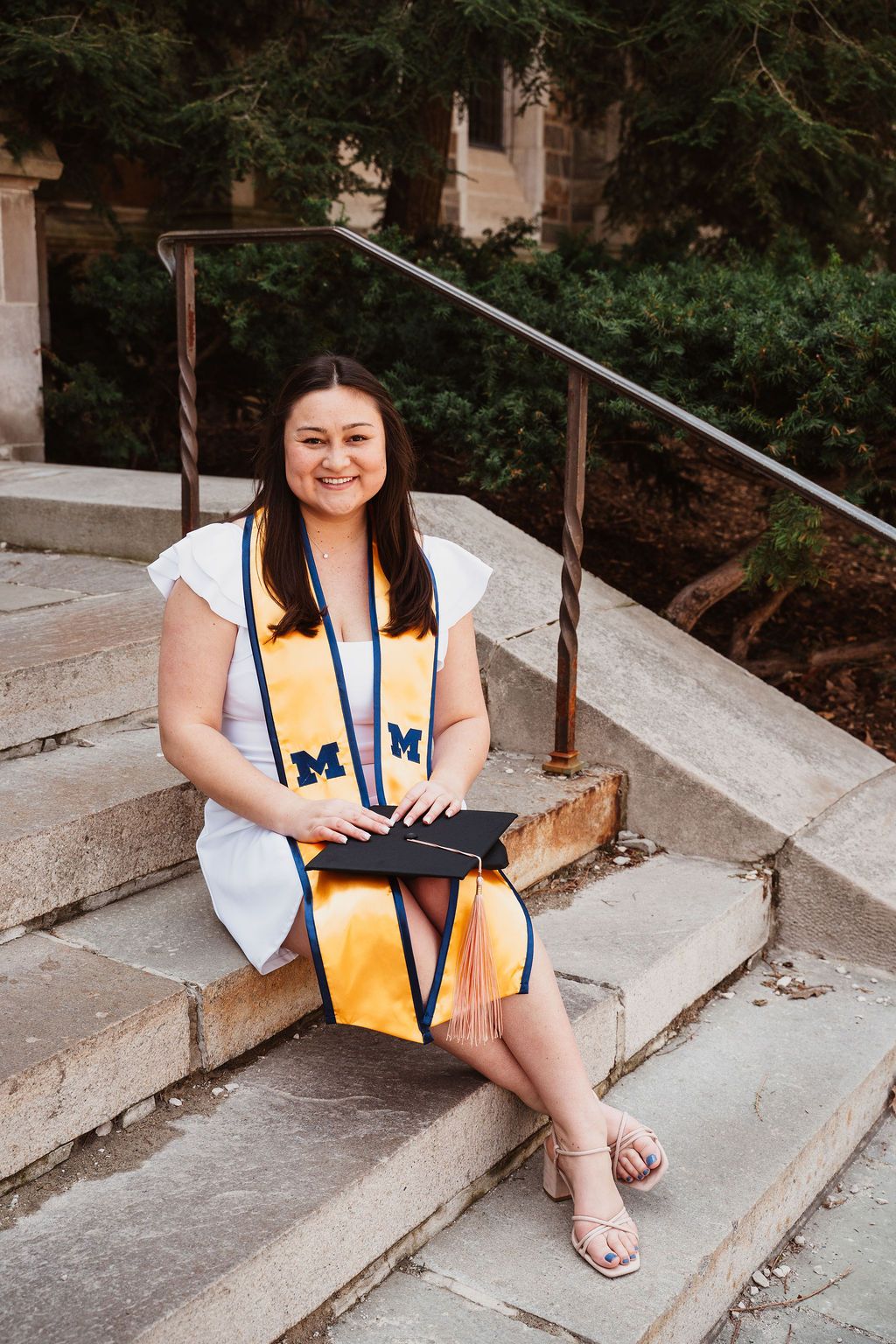 University of Michigan graduate sitting on stone steps holding cap wearing maize and blue stole.