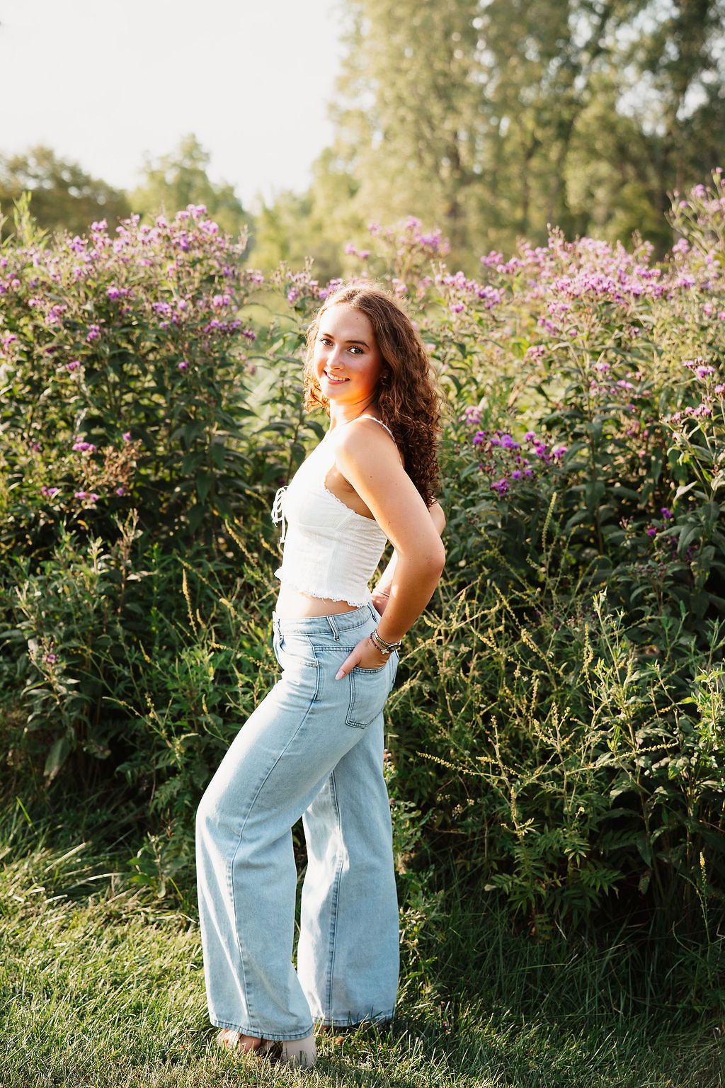 A person in a white top and light denim overalls poses in front of blooming pink wildflowers at sunset.
