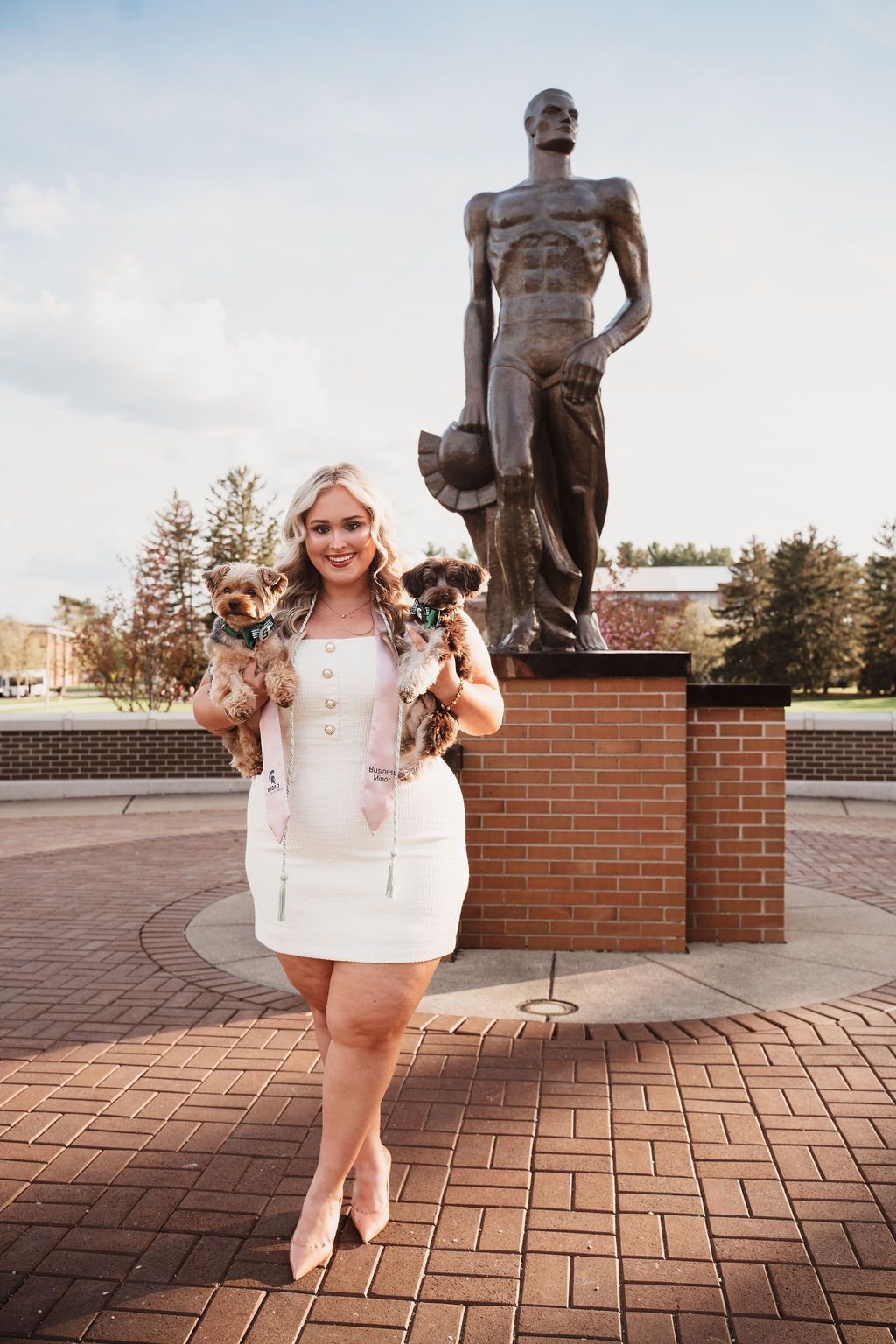 Young woman in white dress holding two small dogs in front of a large bronze Spartan statue on a brick plaza.