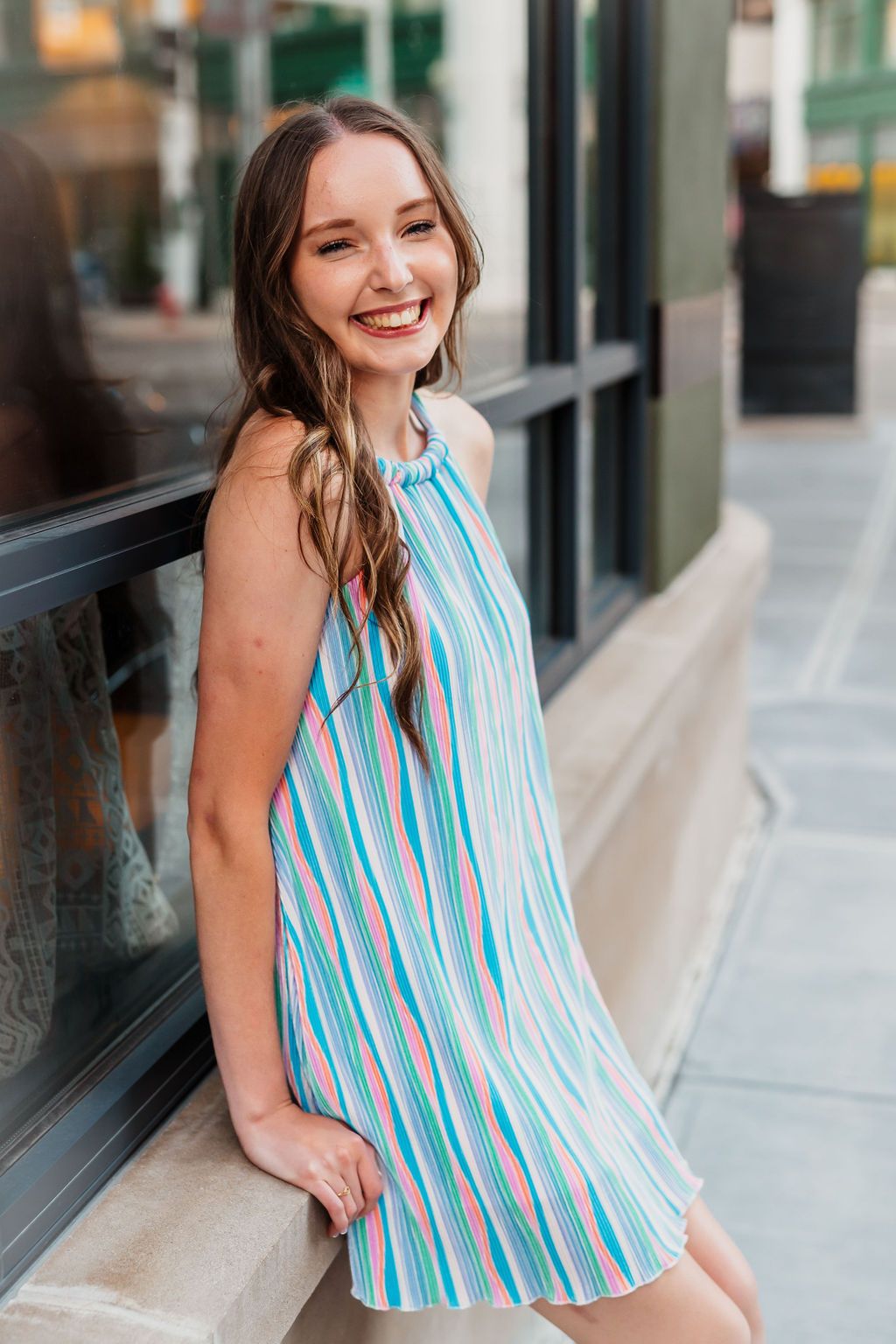 Young woman smiling in a colorful striped halter dress, leaning against a window on an urban sidewalk.