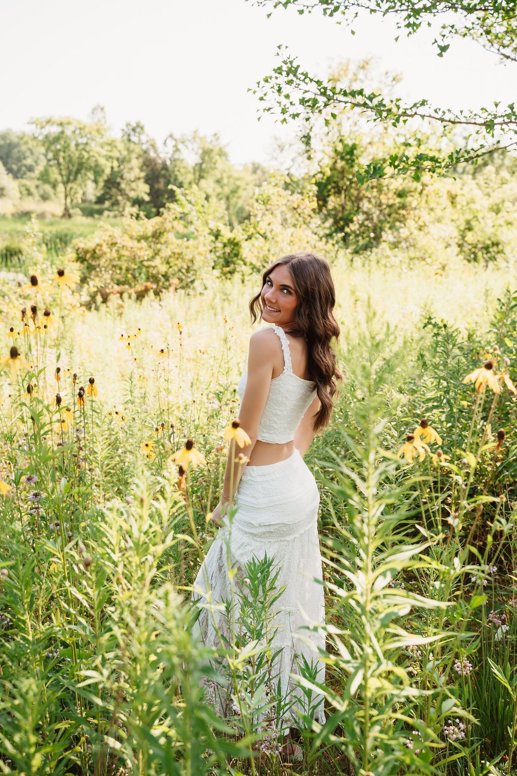 Smiling brunette woman in white crop top and skirt standing among wildflowers in a sunny green meadow.