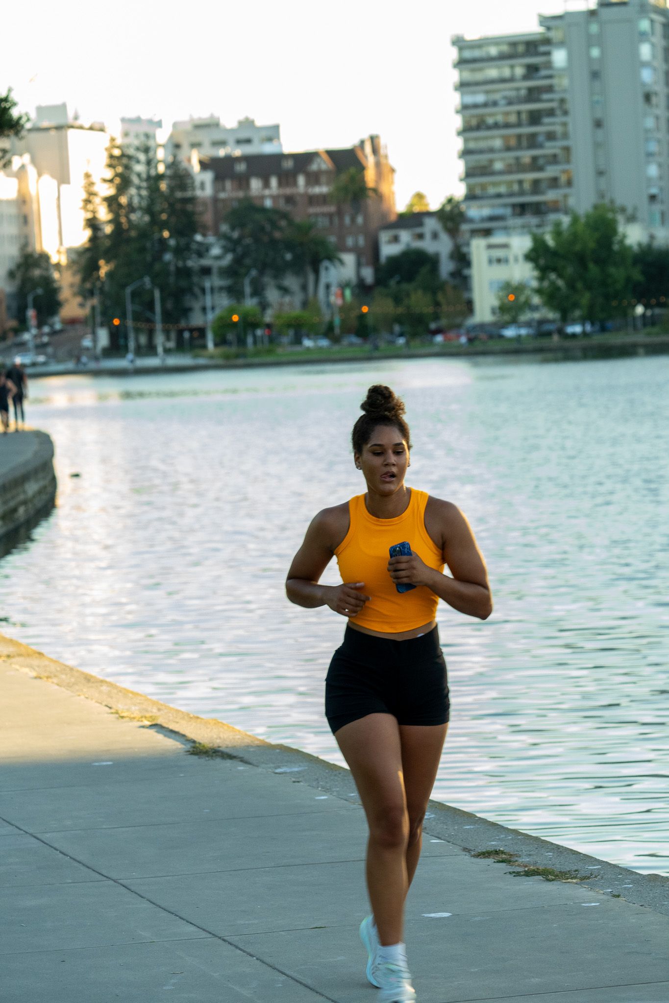 A person in an orange tank top and black shorts jogs along a waterfront path at sunset.