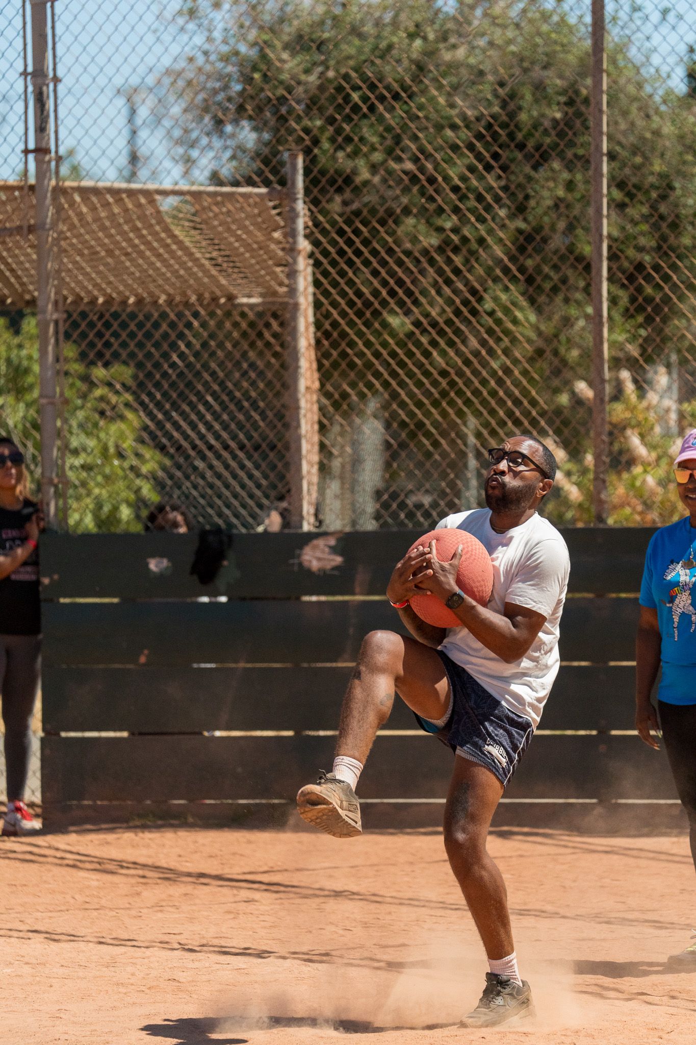Action shot of athletes in motion on a dirt baseball field with chain-link fence backdrop.