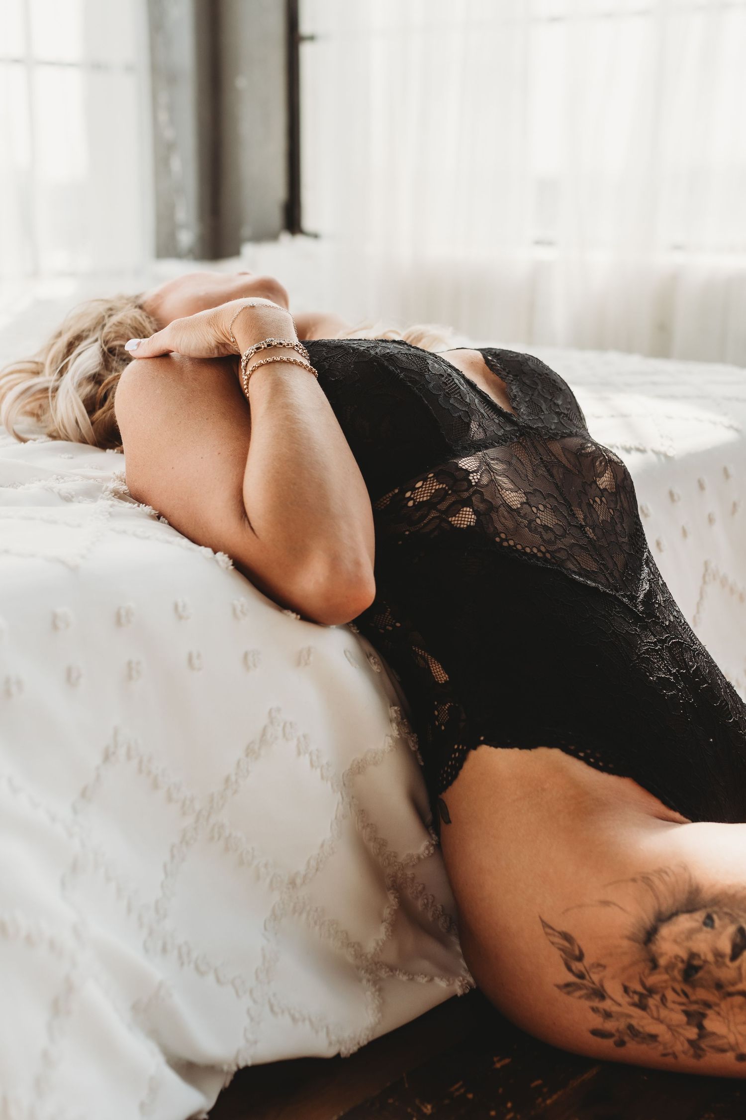 Lowcountry-inspired boudoir portrait featuring a tattooed woman in black lace lingerie on white bedding — authentic and bold.