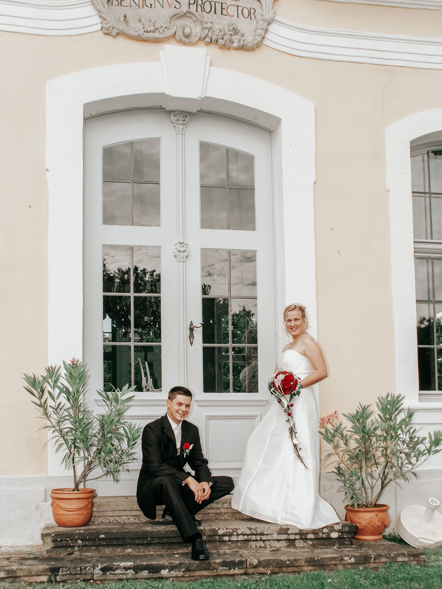A wedding couple poses playfully on a white building exterior with arched windows and potted plants.