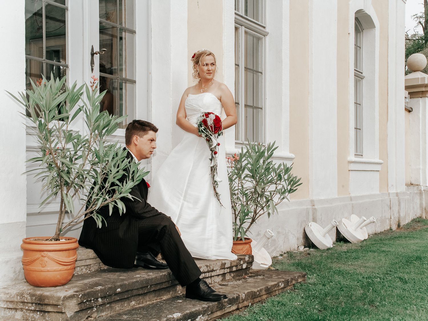 A bride in a white wedding dress and groom in black suit sit on stone steps of an elegant building with potted plants.