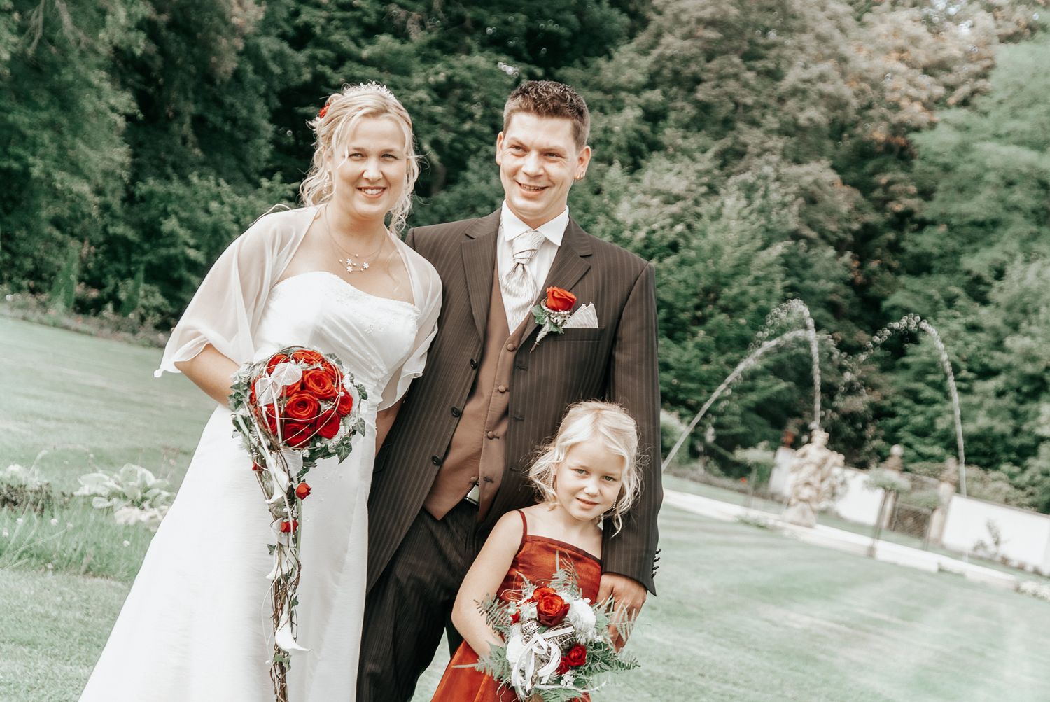 A wedding party poses outdoors in formal attire with red floral bouquets against a green natural backdrop.